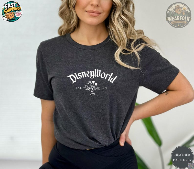A woman wearing a heather dark grey tee with white "DisneyWorld EST 1971" text and Mickey Mouse graphic on the chest.