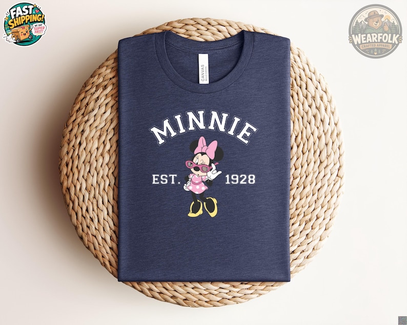 Flat lay of a navy blue t-shirt with Minnie Mouse graphic wearing pink sunglasses, bold white arched text reading "MINNIE" and "EST. 1928" on the chest, displayed folded on a wicker surface.