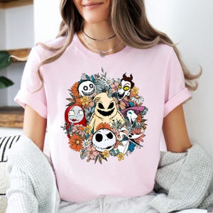 A woman wearing a light pink tee with a colorful floral design featuring smiling skeletal characters from a Halloween movie surrounded by orange and yellow flowers.