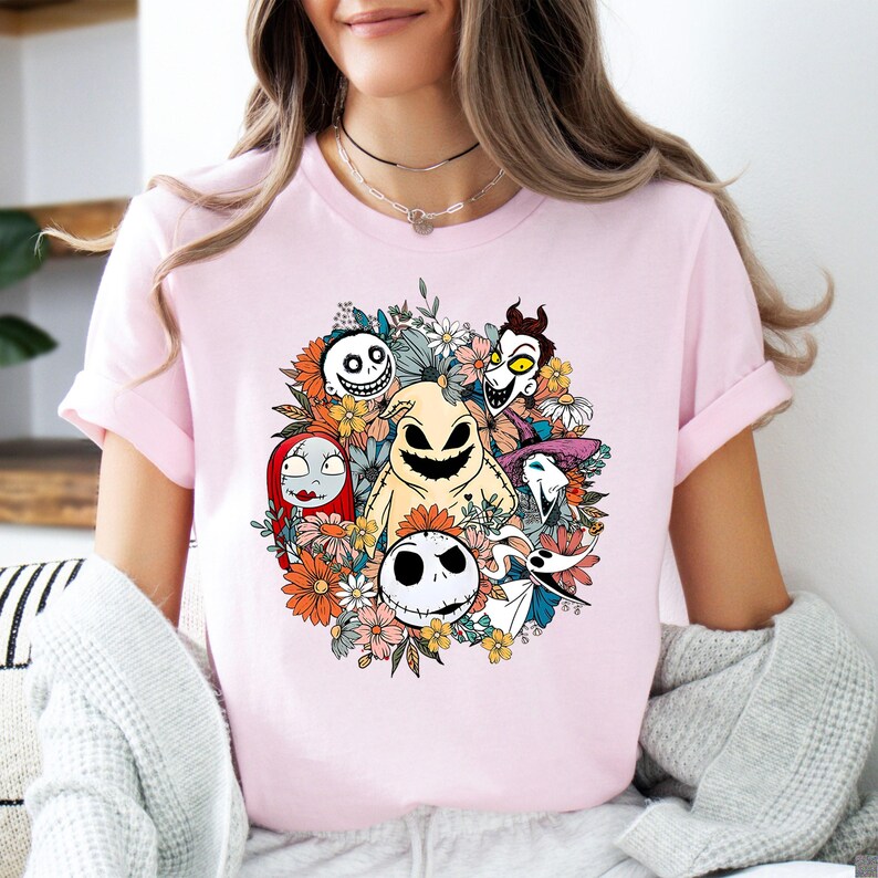 A woman wearing a light pink tee with a colorful floral design featuring smiling skeletal characters from a Halloween movie surrounded by orange and yellow flowers.