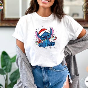 A woman wearing a white Bella Canvas tee with a blue alien character wearing a red Santa hat and holding a candy cane surrounded by snowflakes.