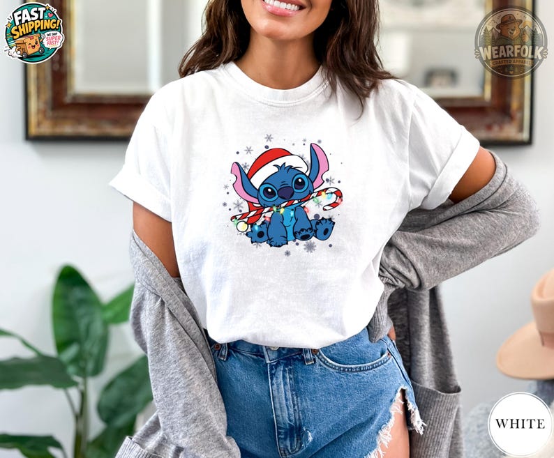 A woman wearing a white Bella Canvas tee with a blue alien character wearing a red Santa hat and holding a candy cane surrounded by snowflakes.
