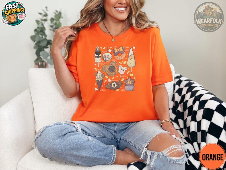 A woman wearing a bright orange tee with a Halloween-themed graphic collage featuring candy, skulls, pumpkins, ghost, and autumn icons in tan and gray.