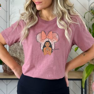 A woman wearing a heather mauve tee with an illustration of Moana's face wearing a polka dot bow and the text "Moana" below.