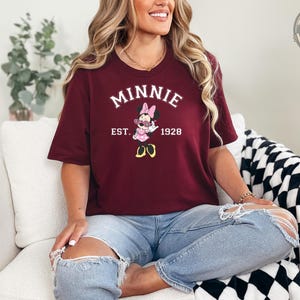 A woman wearing a maroon t-shirt featuring Minnie Mouse in sunglasses with bold white text reading "MINNIE EST. 1928." She's seated on a white chair wearing ripped jeans, smiling.