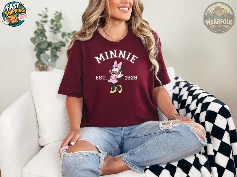 A woman wearing a maroon t-shirt featuring Minnie Mouse in sunglasses with bold white text reading "MINNIE EST. 1928." She's seated on a white chair wearing ripped jeans, smiling.