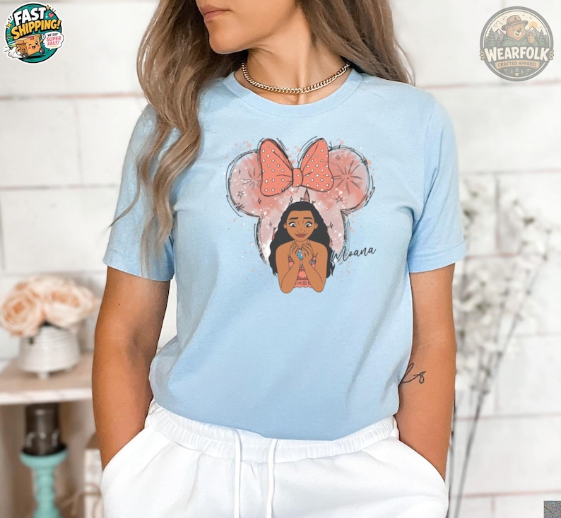 A woman wearing a light blue tee with an illustration of a girl with long black hair inside a mouse head with a red polka dot bow and the name "Moana" below.