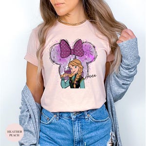 A woman wearing a light pink tee with an illustration of Anna from Frozen holding a present under a purple polka dot bow with sparkles and the name "Anna" written on it.