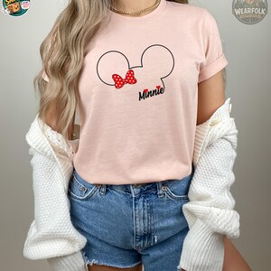 A woman wearing a light pink Bella Canvas tee with Mickey Mouse ears outline, a red polka-dot bow, and "Minnie" text.