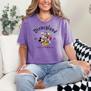 A woman wearing a purple Bella Canvas tee with a Disneyland California graphic featuring Mickey Mouse, Goofy, and other Disney characters with "EST 1955" text.