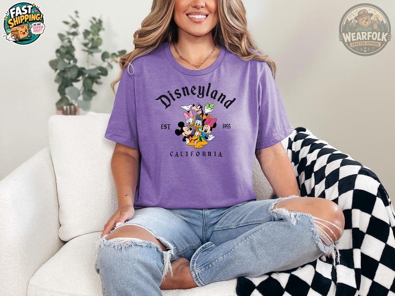 A woman wearing a purple Bella Canvas tee with a Disneyland California graphic featuring Mickey Mouse, Goofy, and other Disney characters with "EST 1955" text.