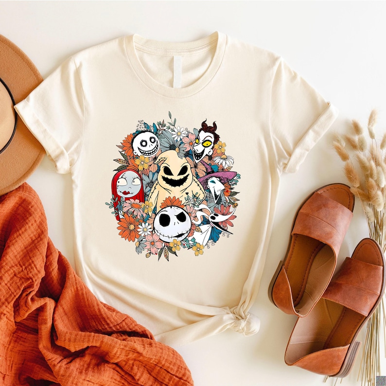 A cream tee with a colorful floral arrangement featuring skeletal characters from a popular Halloween movie in the center of the design.