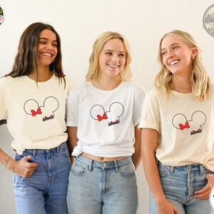 Three women wearing cream and white tees with Minnie Mouse head graphic featuring a red polka dot bow and "Minnie" text.