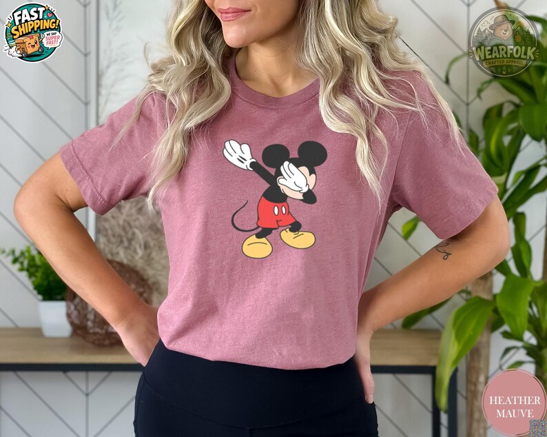 A woman wearing a heather mauve tee with a classic Mickey Mouse graphic showing the character in his iconic red shorts and yellow shoes with white gloves.