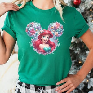 A woman wearing a kelly green tee with a Disney Princess graphic featuring Ariel with red hair inside a floral Mickey ear frame with colorful flowers and sea creatures.