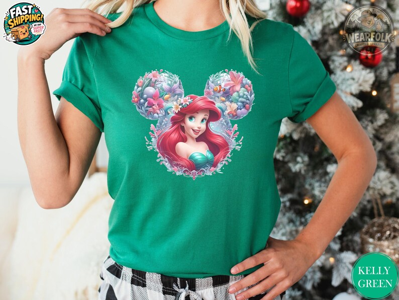A woman wearing a kelly green tee with a Disney Princess graphic featuring Ariel with red hair inside a floral Mickey ear frame with colorful flowers and sea creatures.