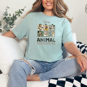 A woman wearing a light blue t-shirt featuring Disney characters in safari outfits arranged in a grid, with bold text reading "ANIMAL KINGDOM" below the characters.