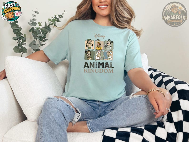 A woman wearing a light blue t-shirt featuring Disney characters in safari outfits arranged in a grid, with bold text reading "ANIMAL KINGDOM" below the characters.
