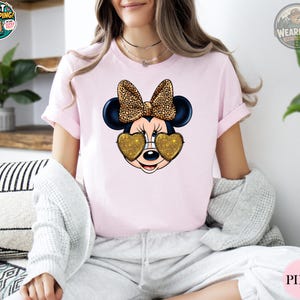 May include: Pink t-shirt featuring a Minnie Mouse graphic. The graphic includes Minnie's face with heart-shaped gold glitter sunglasses and a leopard print bow. The shirt is a short-sleeved crew neck.