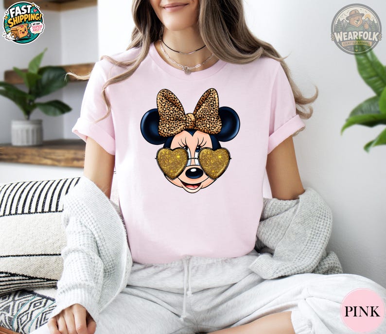 May include: Pink t-shirt featuring a Minnie Mouse graphic. The graphic includes Minnie's face with heart-shaped gold glitter sunglasses and a leopard print bow. The shirt is a short-sleeved crew neck.