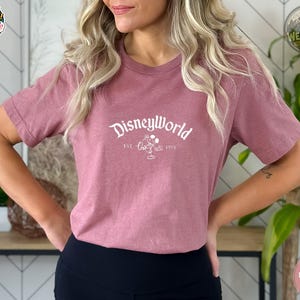 A woman wearing a heather mauve tee with white "DisneyWorld EST 1971" text and Mickey silhouette graphic on the chest.