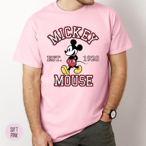 A man wearing a soft pink tee with a classic Mickey Mouse graphic and text reading "Mickey Est. 1928 Mouse" in red and white lettering.