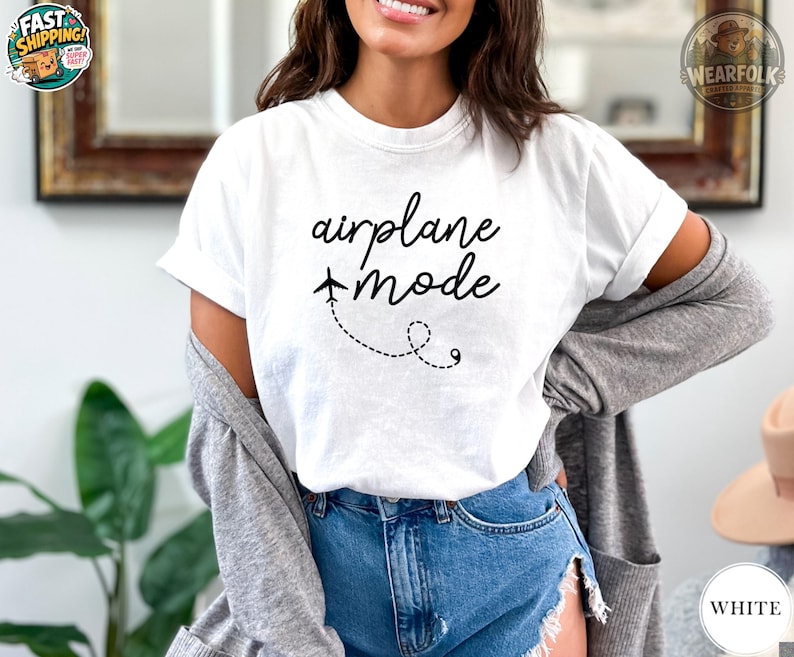 A woman wearing a white t-shirt that reads "airplane mode" in black cursive script with a small airplane and dashed loop design. She is styled with ripped jeans and a grey cardigan.