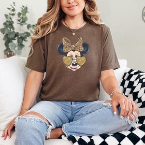 A woman wearing a brown t-shirt featuring Minnie Mouse with a leopard print bow and gold glitter heart-shaped sunglasses, paired with ripped jeans, seated on a white couch.