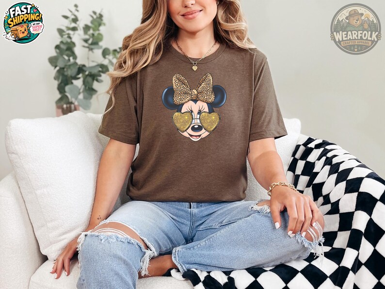 A woman wearing a brown t-shirt featuring Minnie Mouse with a leopard print bow and gold glitter heart-shaped sunglasses, paired with ripped jeans, seated on a white couch.