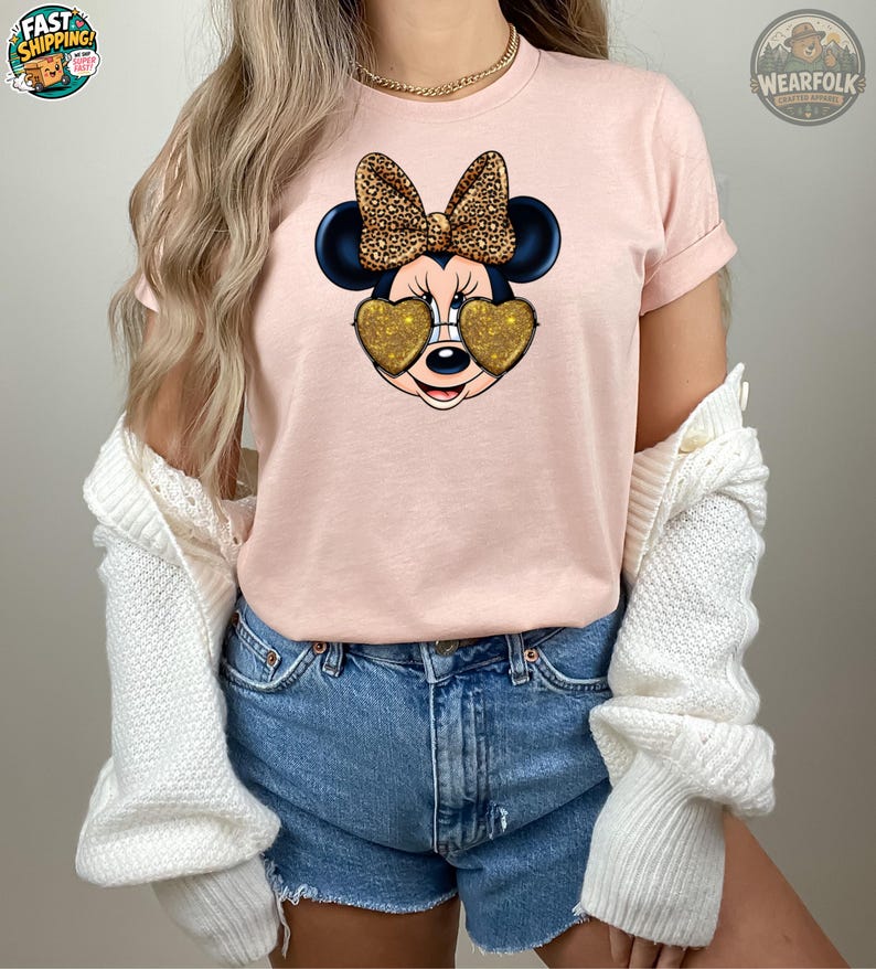 A woman wearing a light pink t-shirt featuring Minnie Mouse with gold glitter heart-shaped sunglasses and a leopard print bow, styled with denim shorts and a white knit cardigan.