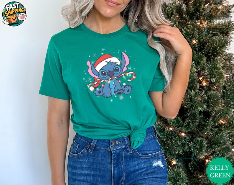 Woman wearing a kelly green tee with a blue alien character wearing a Santa hat and holding a candy cane surrounded by snowflakes.