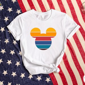 White tee with a colorful Mickey Mouse head silhouette featuring orange, red, navy, teal, and brown horizontal stripes.