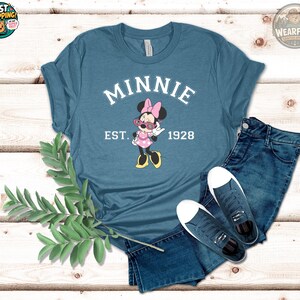 Flat lay of a steel blue t-shirt with Minnie Mouse graphic wearing pink sunglasses, bold white arched text reading "MINNIE" above and "EST. 1928" below, styled with denim shorts and sneakers on white wood.