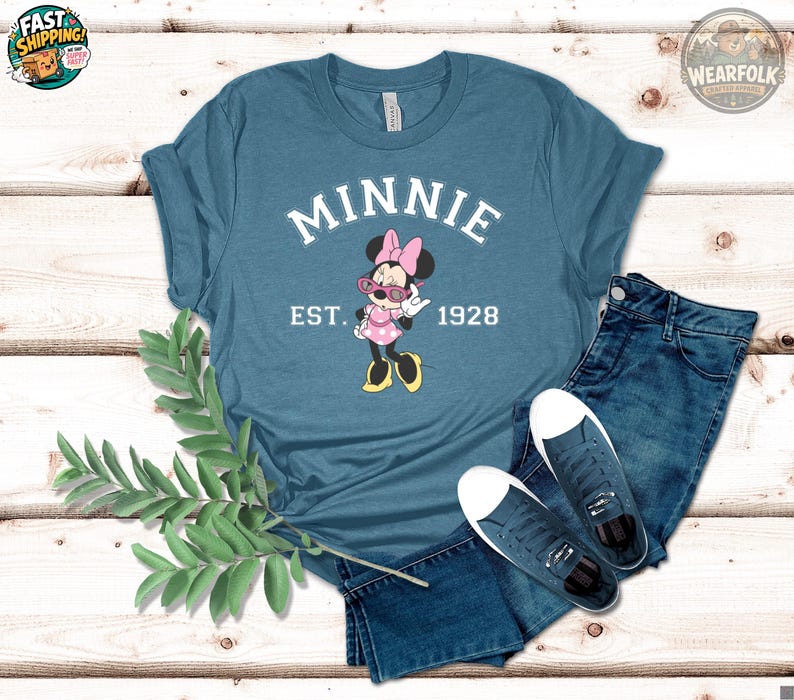 Flat lay of a steel blue t-shirt with Minnie Mouse graphic wearing pink sunglasses, bold white arched text reading "MINNIE" above and "EST. 1928" below, styled with denim shorts and sneakers on white wood.
