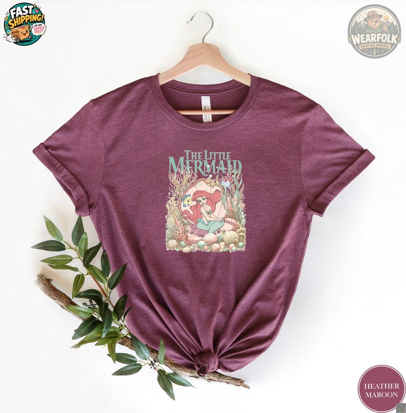 Heather maroon tee featuring "The Little Mermaid" title with Ariel swimming among coral and seaweed in vintage illustration style.