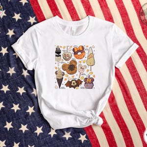 White Bella Canvas tee with a graphic featuring Halloween-themed ice cream, donuts, candy corn, and character illustrations with labels scattered across the chest.