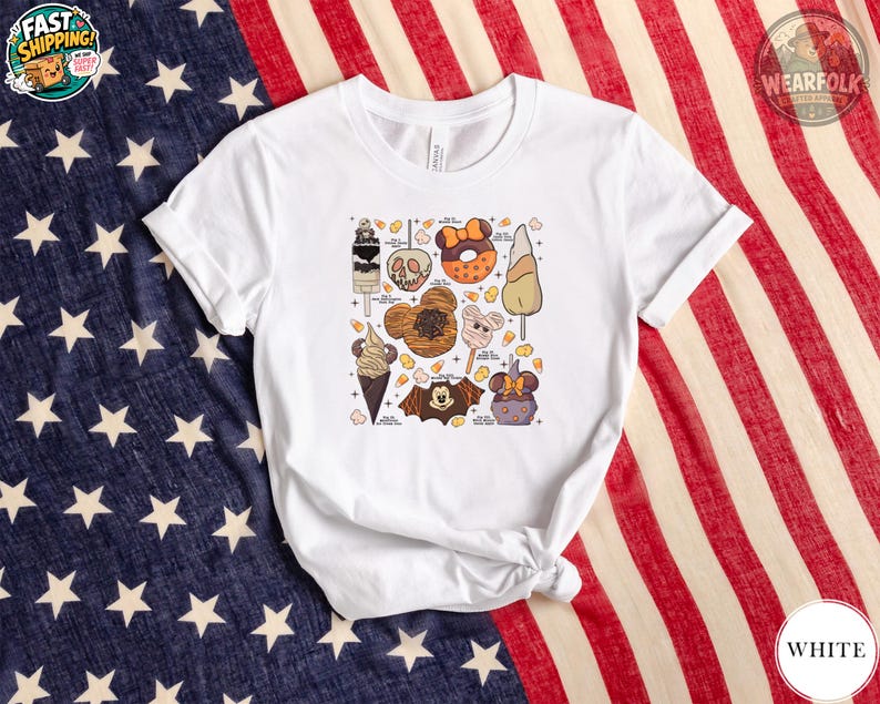 White Bella Canvas tee with a graphic featuring Halloween-themed ice cream, donuts, candy corn, and character illustrations with labels scattered across the chest.