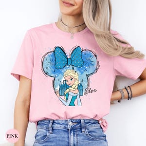 A woman wearing a pink tee with an Elsa character illustration inside a blue polka dot bow shape and castle, with the name "Elsa" written below.