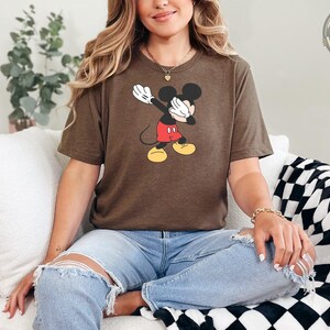 A woman wearing a brown Bella Canvas tee with a vintage Mickey Mouse graphic showing the character with white gloves raised up, red shorts, and yellow shoes.