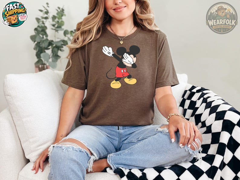 A woman wearing a brown Bella Canvas tee with a vintage Mickey Mouse graphic showing the character with white gloves raised up, red shorts, and yellow shoes.