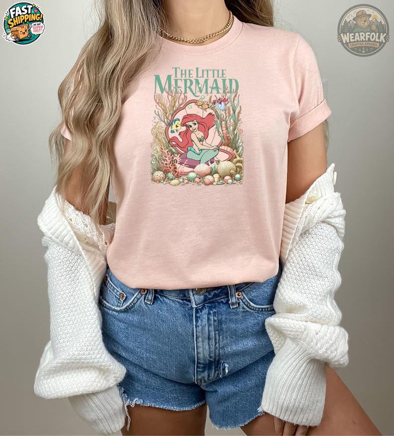 Puede incluir: Una camiseta rosa claro con un dise&ntilde;o de Ariel de La Sirenita. El dise&ntilde;o incluye el texto "The Little Mermaid" en verde, con Ariel en una concha rodeada de plantas acu&aacute;ticas y conchas marinas. La camiseta es usada.