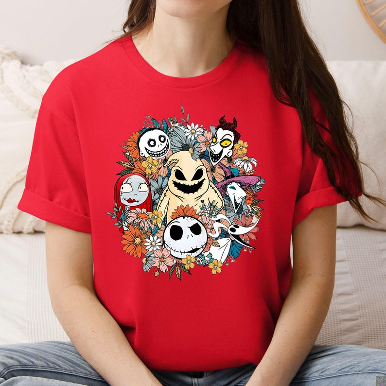 A woman wearing a red tee with a floral graphic featuring cartoon skeleton and ghost characters arranged in a circular wreath design.