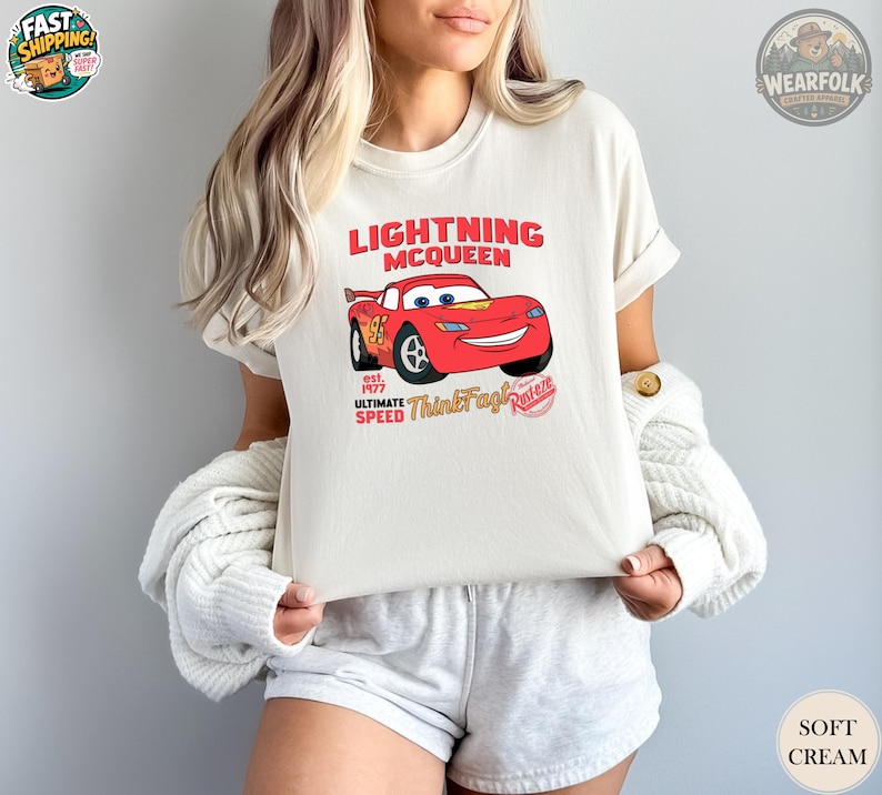 Woman wearing a cream tee with a red Lightning McQueen car graphic and text reading "Lightning McQueen est. 1977 Ultimate Speed ThinkFast Piston.