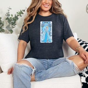 A woman wearing a heather dark grey tee with a tarot-style graphic of Elsa in a blue gown surrounded by snowflake borders and text reading "Elsa".