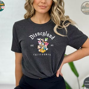 A woman wearing a heather dark grey Bella Canvas tee with Disneyland California EST 1955 graphic featuring Mickey, Minnie, Donald Duck and Pluto.