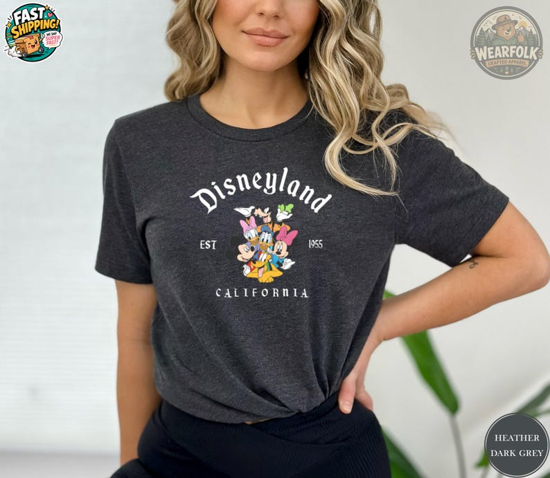 A woman wearing a heather dark grey Bella Canvas tee with Disneyland California EST 1955 graphic featuring Mickey, Minnie, Donald Duck and Pluto.