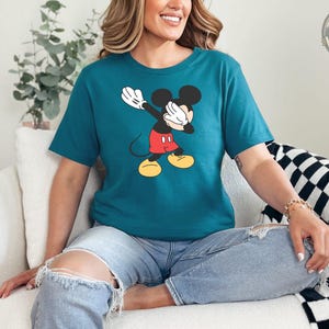 A woman wearing a teal tee with a classic Mickey Mouse graphic featuring white gloves, black ears, and red shorts with yellow shoes.