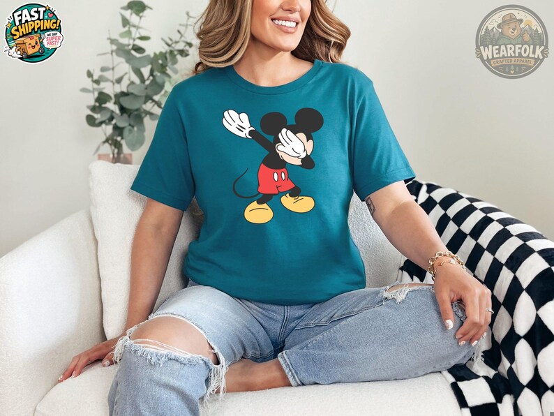 A woman wearing a teal tee with a classic Mickey Mouse graphic featuring white gloves, black ears, and red shorts with yellow shoes.