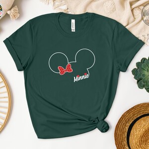 A woman wearing a forest green Bella Canvas tee with a Minnie Mouse head outline and red polka dot bow graphic that says "Minnie" below.