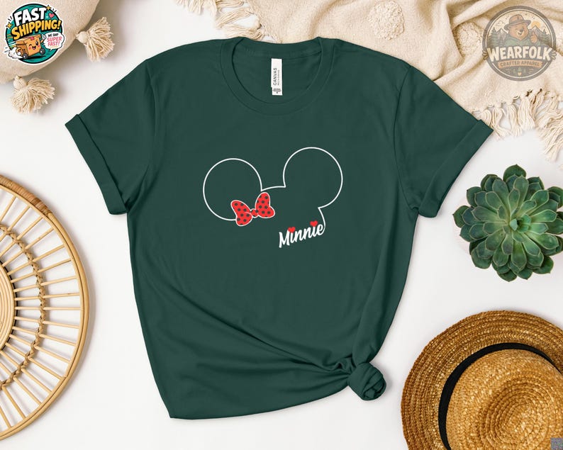 A woman wearing a forest green Bella Canvas tee with a Minnie Mouse head outline and red polka dot bow graphic that says "Minnie" below.